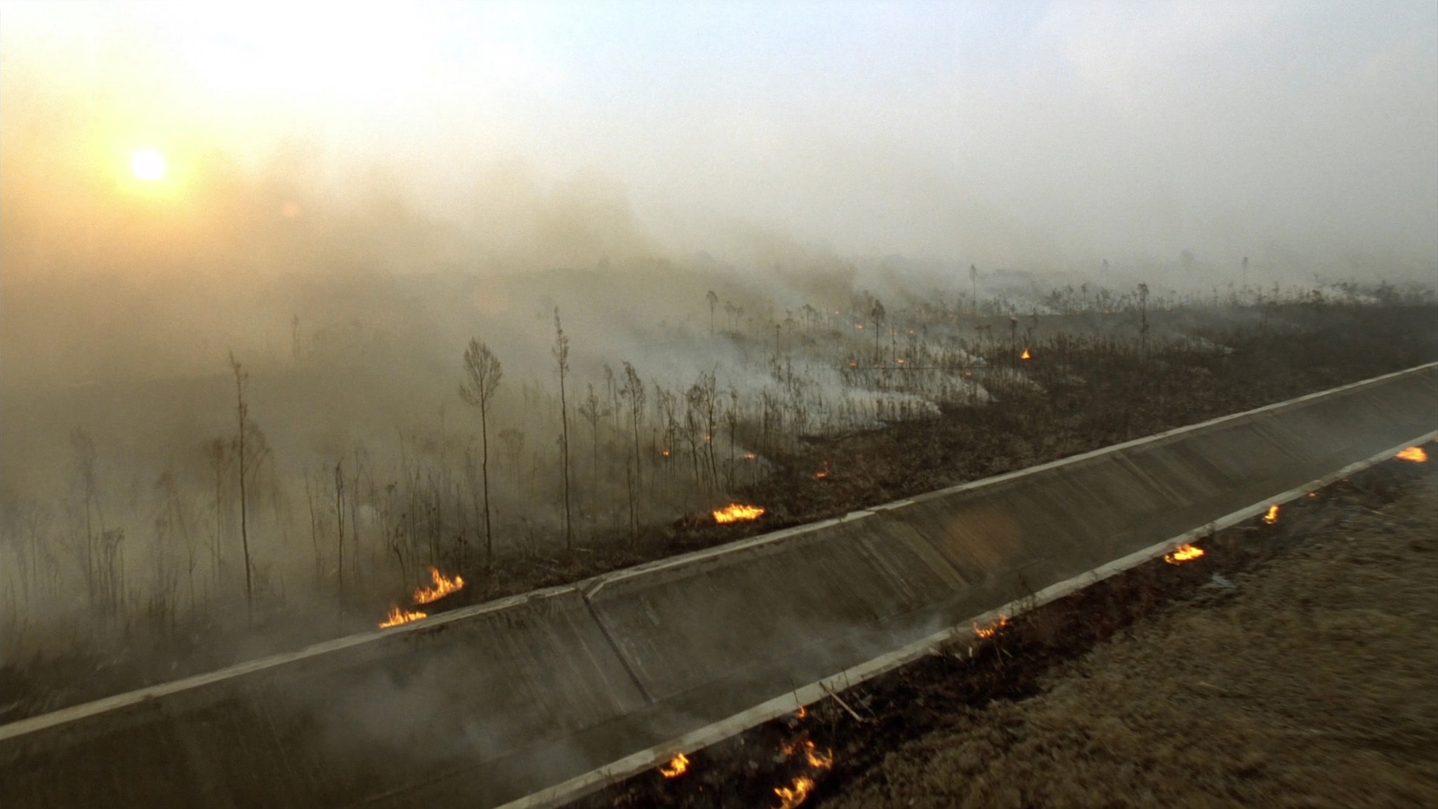 A concrete-lined drainage canal running diagonally through degraded peatland, with lines of low flame burning along both banks and smoke drifting across the landscape.